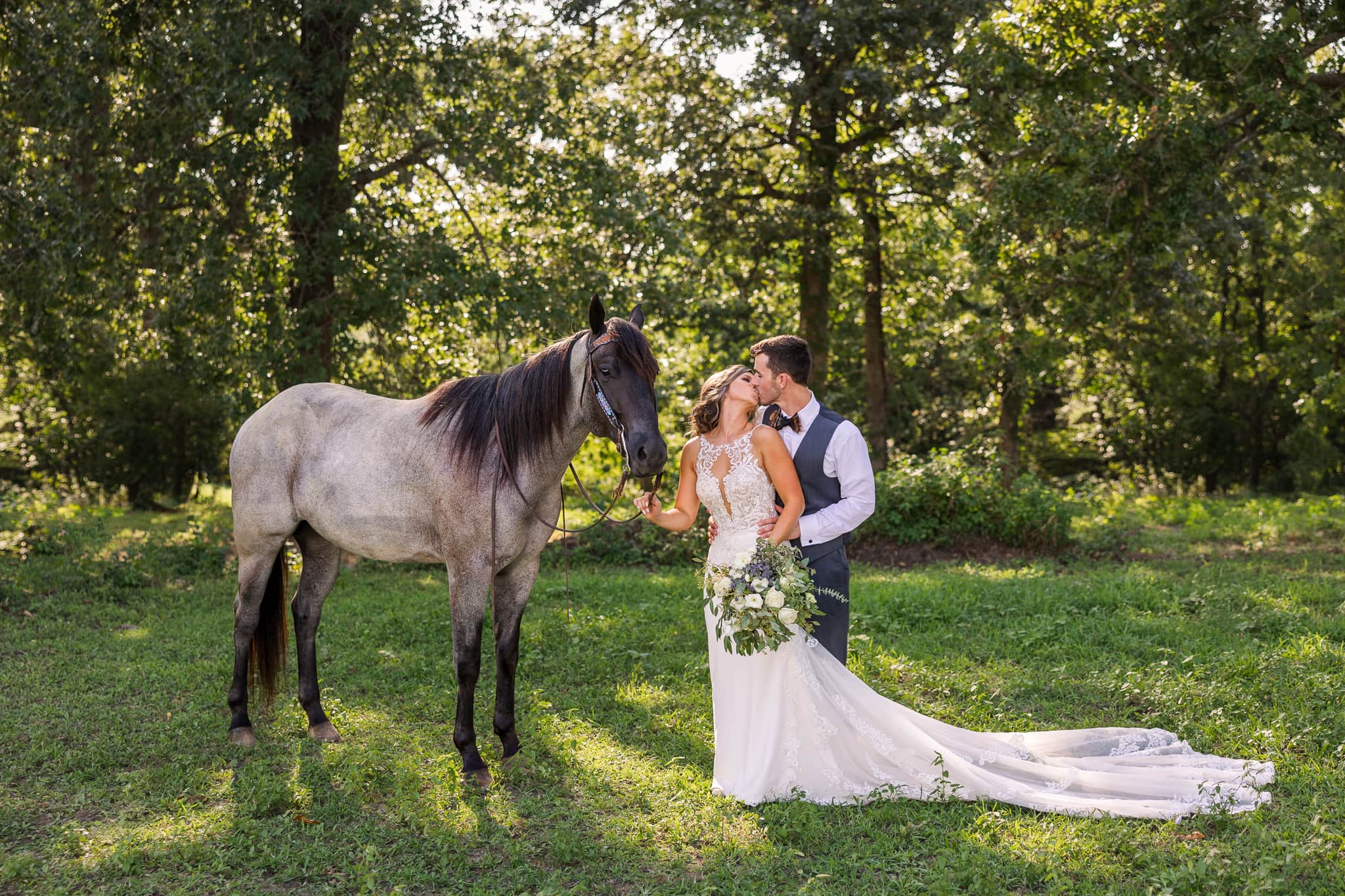 Outdoors in front of stand of trees, with the sun filtering through behind, stands a bride and groom, sharing a kiss with their beloved horse beside them.
