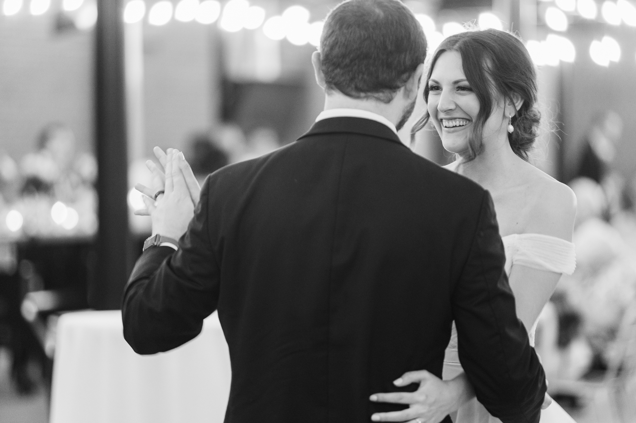 A black & white photo of a bride and groom dancing with twinkle lights in the background